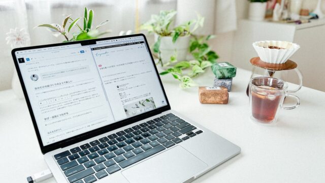 白いデスクの上に置かれたノートパソコンが開かれており、画面にはブログの編集画面が表示されています。パソコンの右側にはガラスのカップに注がれたコーヒーが置かれており、その隣にはコーヒーのハンドドリップに使う道具があります。す。背景には観葉植物が飾られています。
